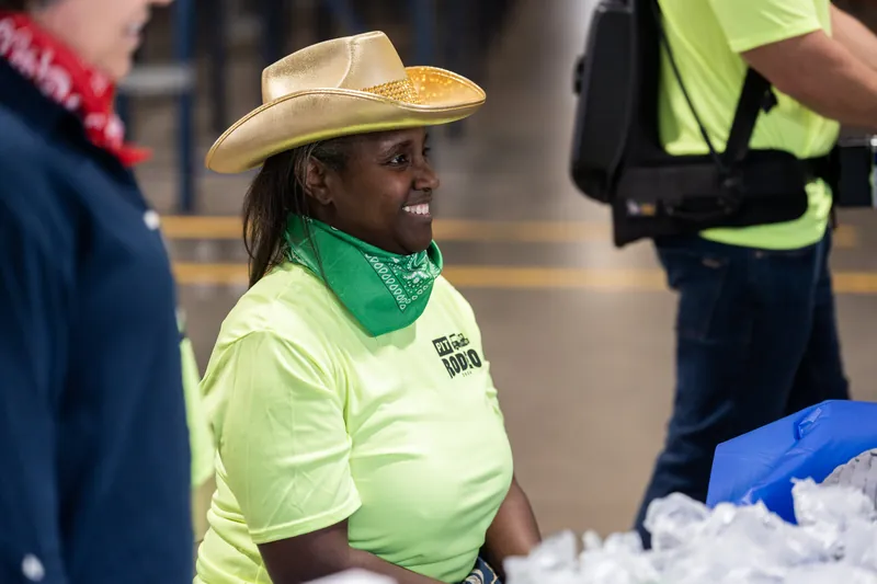 A smiling person wearing a tan cowboy hat, lime-green shirt, and green bandana stands among event attendees indoors.