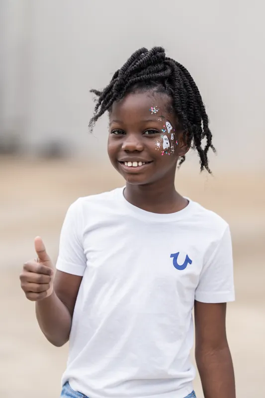 Child in a white t-shirt with a blue horseshoe logo gives a thumbs-up while wearing face glitter at the beach rodeo event.
