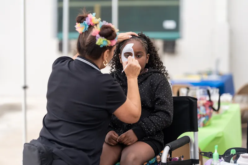 Two people sit together as one applies face paint to the other's cheek at an indoor rodeo activity station.