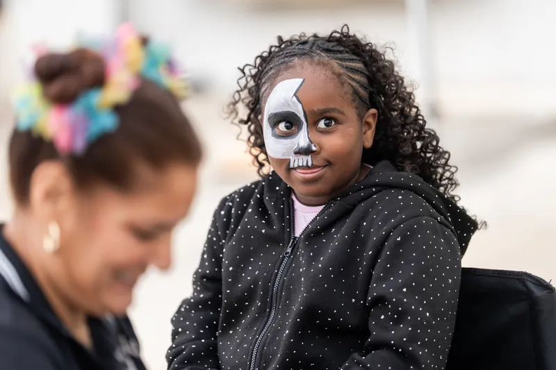 Child with face paint designed as a skull smiles while wearing a black polka-dot jacket at the rodeo celebration.