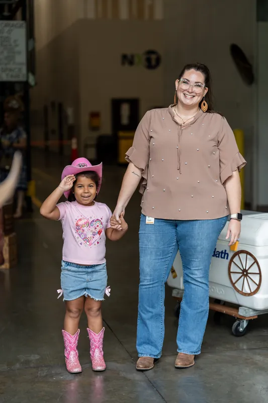 An adult in a tan shirt and child in pink boots hold hands while walking through an indoor venue space.