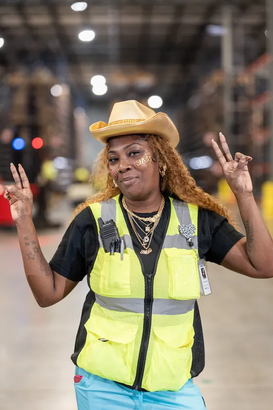 A person wearing a yellow cowboy hat and reflective safety vest makes a peace sign with both hands.