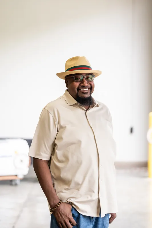 A man in a cream button-up shirt and tan hat smiles while standing indoors at the rodeo event.
