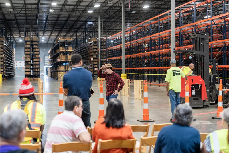 Participants in safety gear and hard hats stand among orange traffic cones near a forklift inside a warehouse during a pit rodeo event.