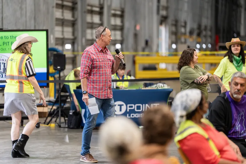 A person in a pink plaid shirt speaks to a crowd while colleagues in yellow safety gear stand nearby at an indoor warehouse venue.
