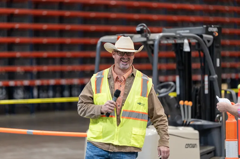 A man wearing a cowboy hat, tan jacket, and neon safety vest stands in front of industrial shelving and a forklift in a warehouse.