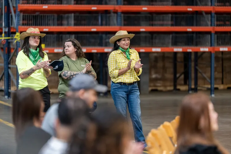 Three people in cowboy hats and western attire address an audience gathered in front of orange and blue industrial racking.