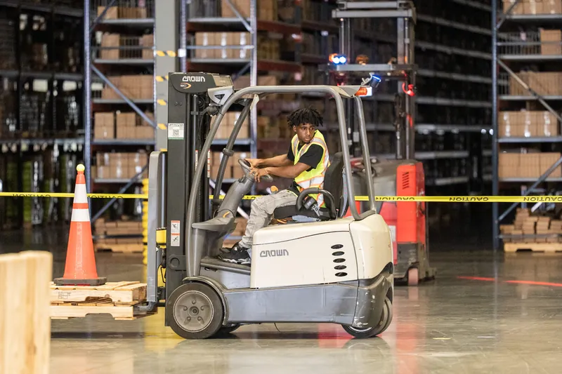 A person operates a small forklift truck inside a warehouse facility surrounded by tall metal shelving and safety cones.