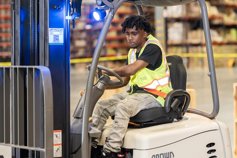 Someone in a black shirt and yellow safety vest sits in the operator's seat of a white forklift, holding the steering wheel inside a warehouse.