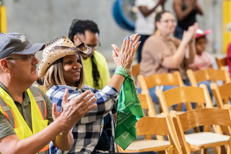 Two people high-five while wearing cowboy hats and safety vests at an indoor rodeo event with wooden bleachers behind them.