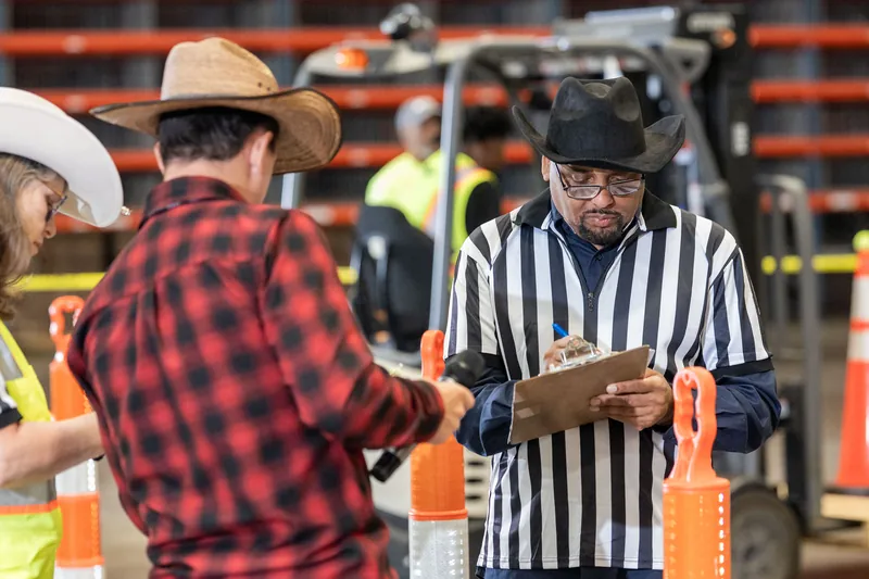 A rodeo official in a black-and-white striped shirt and cowboy hat shakes hands with a participant in a red plaid shirt.