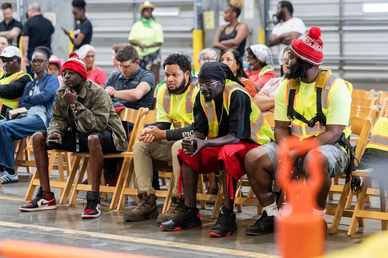 Seated spectators in safety vests and cowboy hats watch the action unfold at the pit rodeo, with an orange obstacle visible in the foreground.