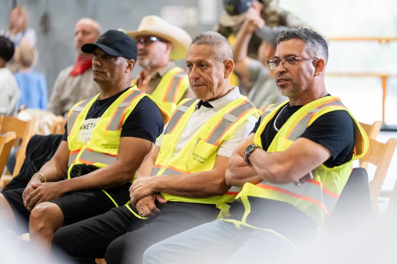 Three men in yellow safety vests sit together on bleachers, observing the event with focused expressions.