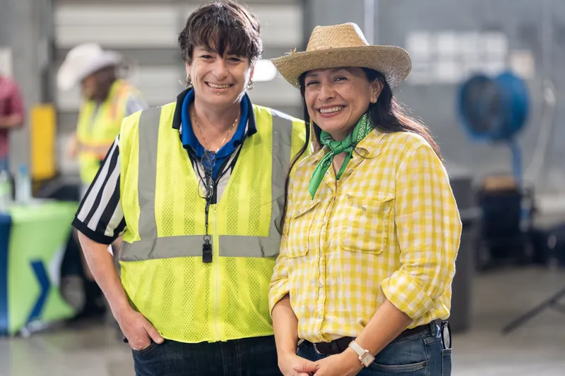 Two event staff members smile side by side—one in a neon safety vest and the other in a yellow gingham shirt and tan cowboy hat.