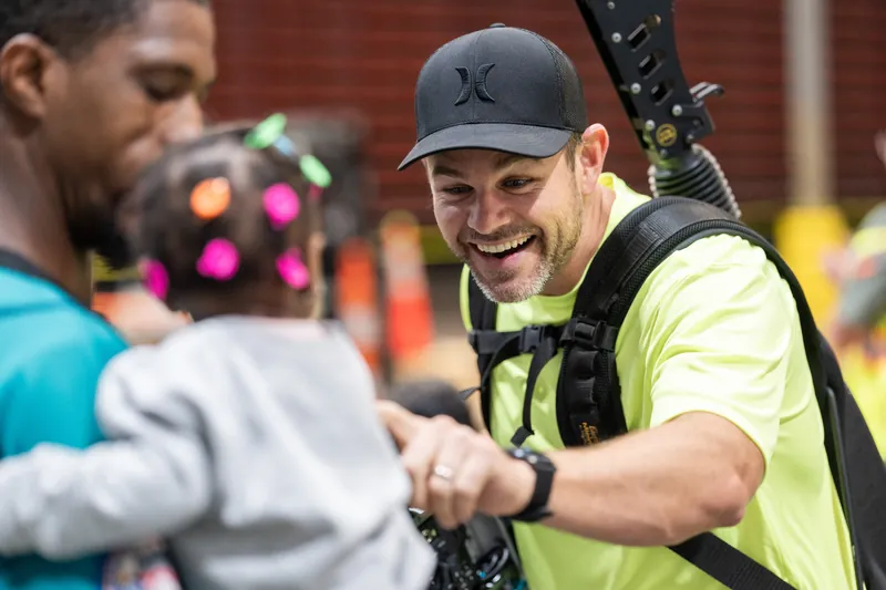 A man in a neon safety vest and dark cap grins while shaking hands with someone in a turquoise shirt during the rodeo activities.