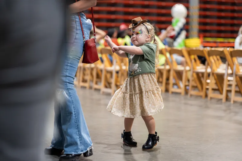 A young child in a patterned dress reaches toward an adult's hand on an indoor rodeo arena floor lined with wooden chairs.