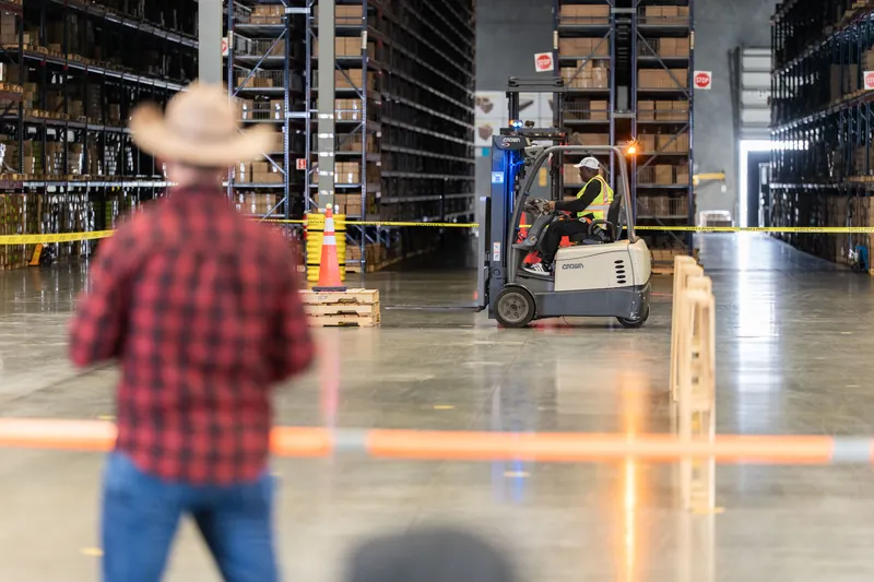 A person in a red plaid shirt stands in an industrial warehouse as a forklift operator navigates stacked wooden pallets.