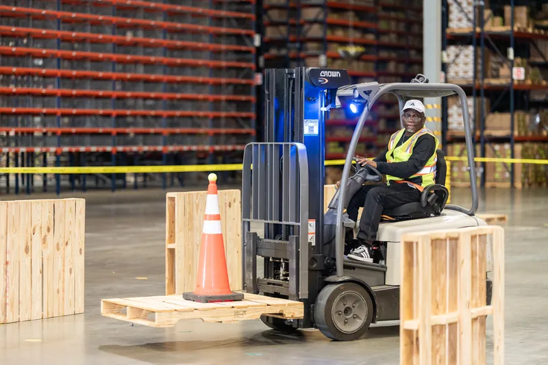 An operator in a yellow safety vest sits in the seat of a red forklift surrounded by wooden pallets and yellow caution tape.