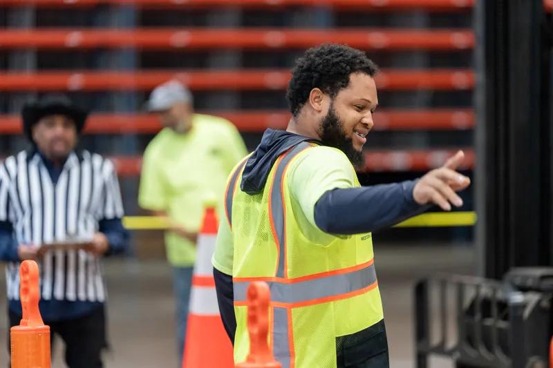 A smiling staff member in a neon yellow safety vest gestures while directing activity at the rodeo event behind him.