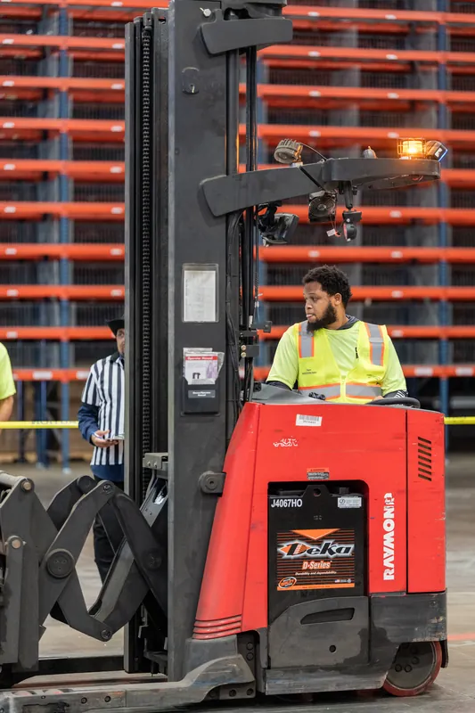 A red forklift with its operator sits against an orange-and-black striped backdrop with safety cones visible in the frame.