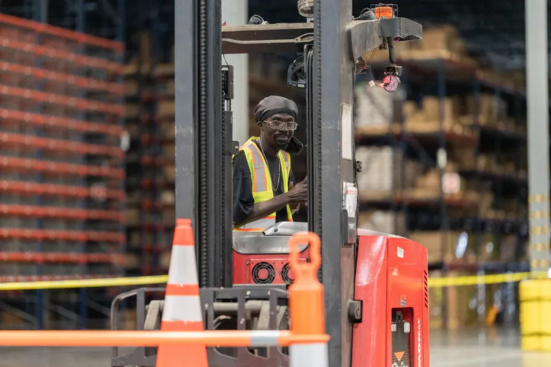 Worker in a yellow safety vest and hard hat sits in the cab of a red forklift surrounded by traffic cones and warehouse shelving.