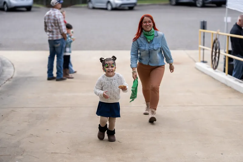 An adult and child walk together on a concrete surface at Jacksonville Beach, St. Augustine, with parked cars visible behind them.