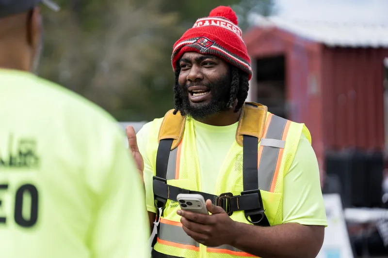 Someone wearing a red beanie and yellow safety vest smiles while holding a clipboard at the rodeo event.