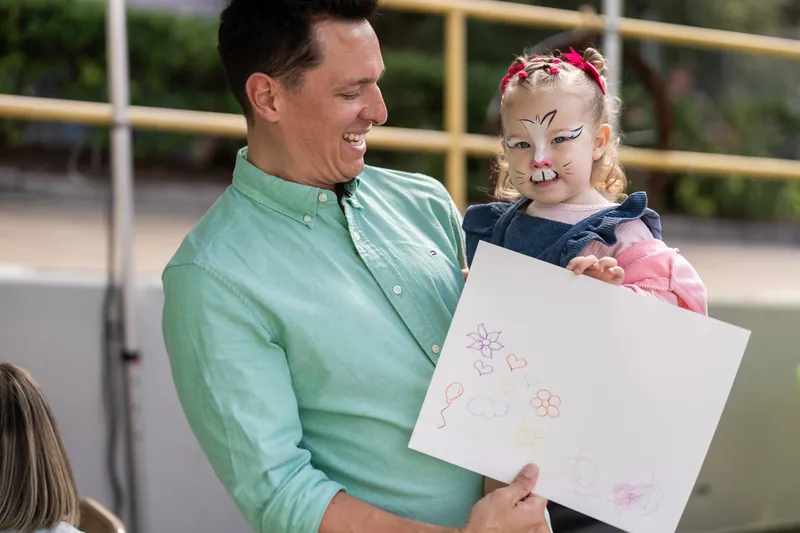 A man in a green shirt holds up a child's drawing while standing with a young child wearing a pink headband.