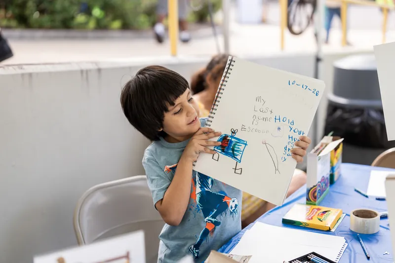 A child at a table concentrates while drawing on a large paper with markers during an activity session.