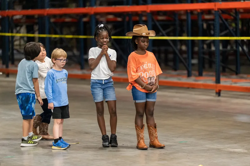 Four children stand together on pavement near a red metal fence railing at the pit rodeo.