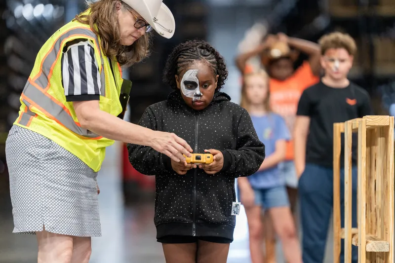 An adult volunteer in a yellow safety vest hands a small toy to a child wearing face paint at the event.
