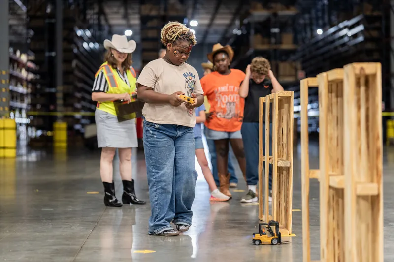 Group of children and adults in safety vests and hard hats walking past wooden pallets indoors.