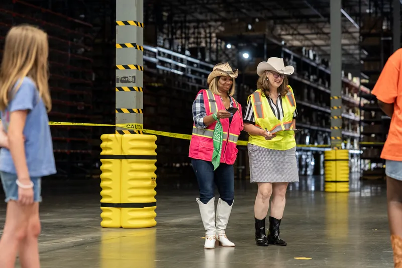 Two people in neon safety gear and cowboy hats standing near yellow barrels and caution tape.
