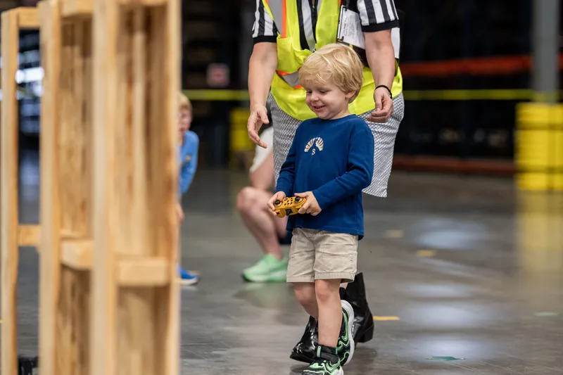 Young child in blue shirt running while holding a toy, guided by an adult in safety vest.
