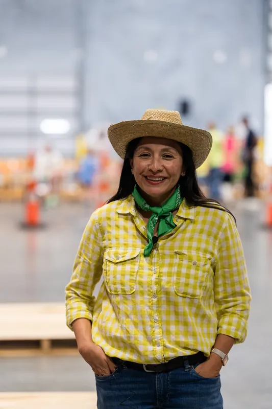Woman in yellow gingham shirt and straw cowboy hat smiling at the camera on an indoor concrete floor.