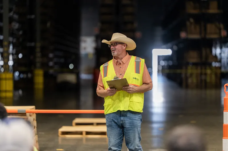 Man in yellow safety vest and tan cowboy hat holding a clipboard inside a warehouse facility.