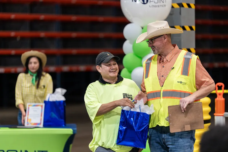 Man in neon vest and cowboy hat hands a blue gift box to another man in matching safety gear at Jacksonville Beach pit rodeo event.