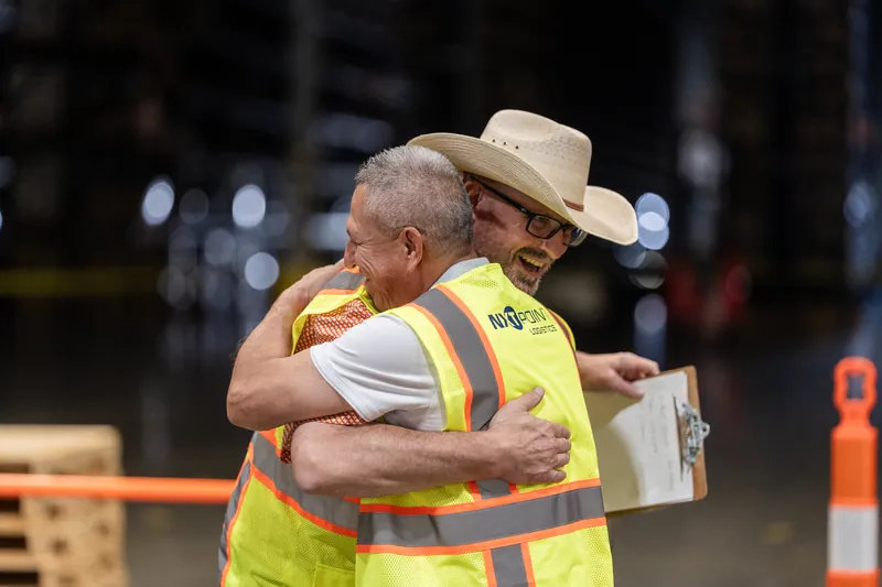 Two people in yellow safety vests embrace warmly, one wearing a tan cowboy hat, during the corporate rodeo celebration.