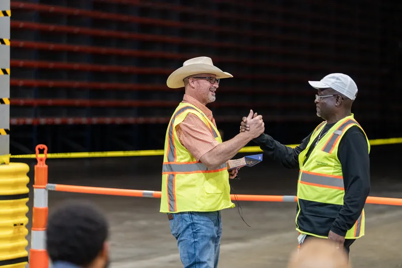 Two event coordinators in fluorescent vests shake hands on the pit rodeo floor, one in a cream cowboy hat.