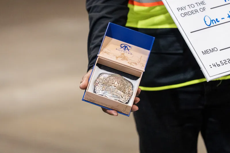 Close-up of hands holding an open wooden box containing a large silver belt buckle, part of the rodeo prize package.
