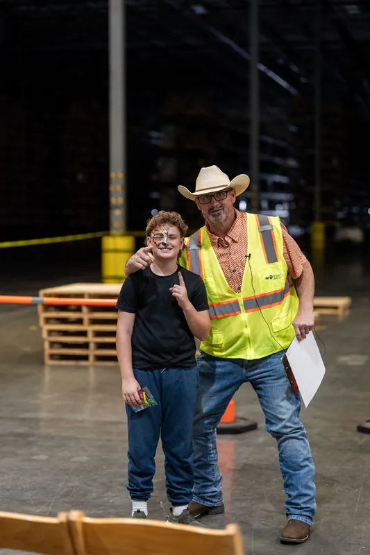 An adult in a yellow safety vest and cowboy hat stands beside a child in glasses and a black shirt, both smiling in an indoor arena.