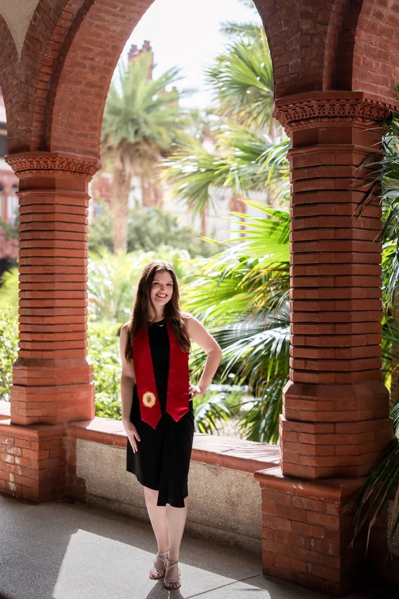 Young woman in black dress and red vest standing under red brick arches with palm trees visible in the background.