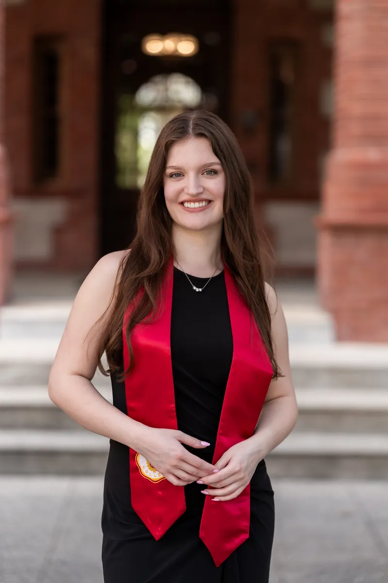 Woman with long dark hair wearing a red and black outfit, smiling at the camera in front of an ornate brick archway.