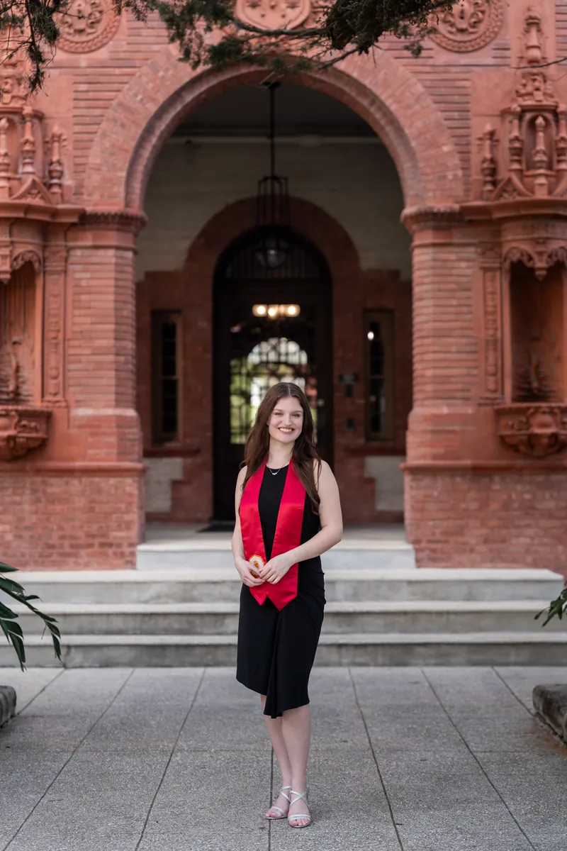 Woman in black dress with red scarf standing alone on brick steps in front of an elaborate red brick building entrance.