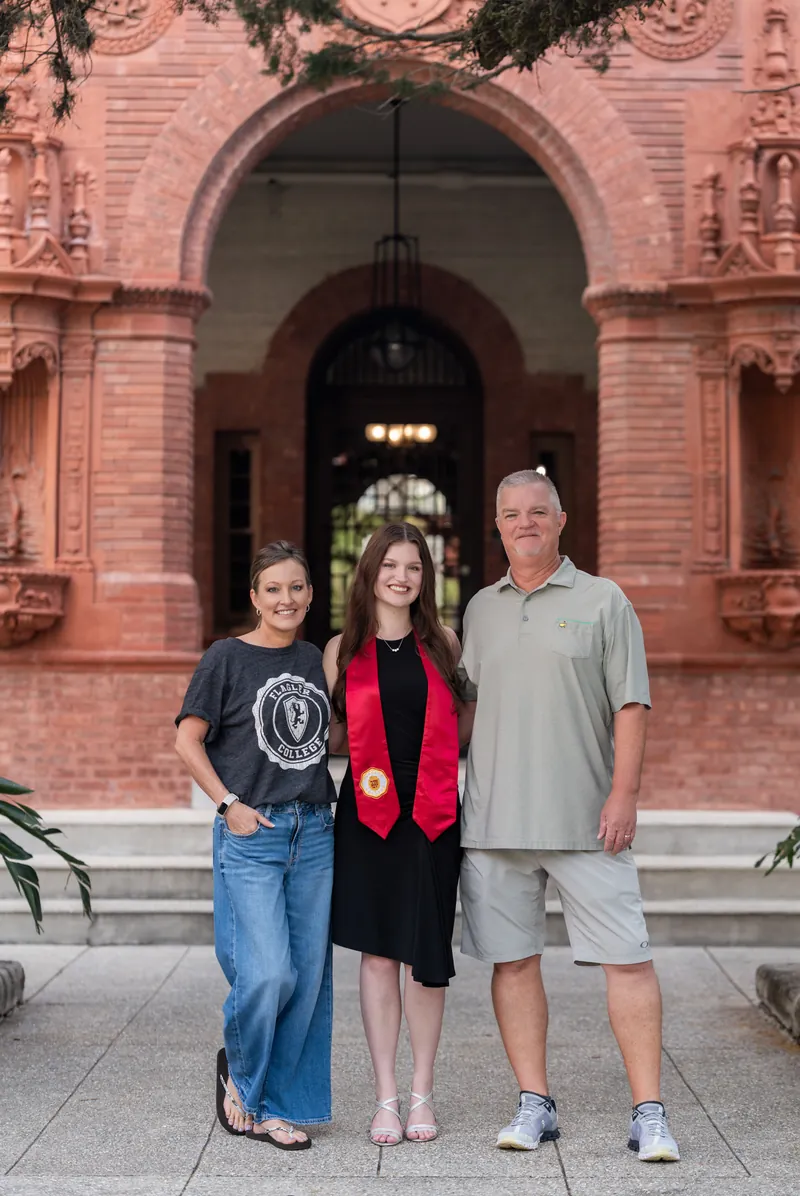 Three people on brick steps at an ornate red brick archway, woman in center wearing red and black, flanked by two adults