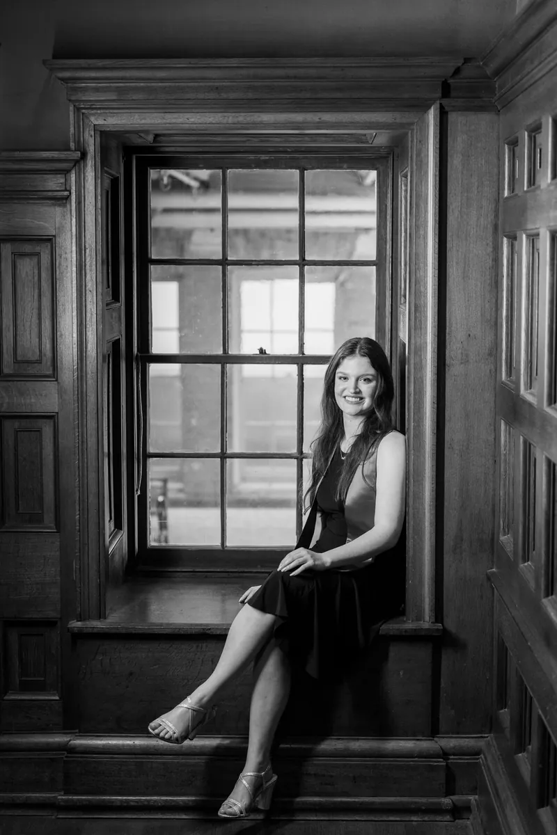 Black and white portrait of woman in dark sleeveless dress sitting by a window with gridded panes, looking toward the camera.