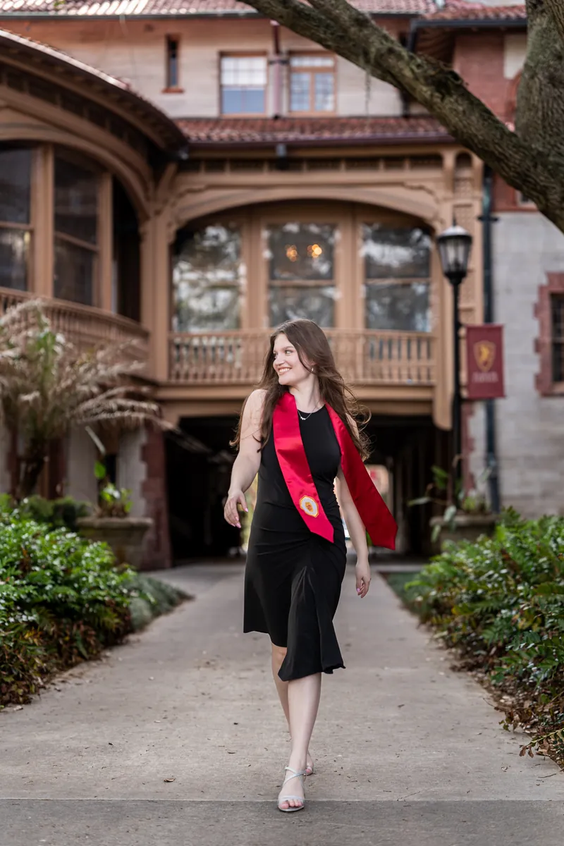 Olivia walks down a garden pathway in a black dress and red scarf, passing manicured hedges with a historic building behind