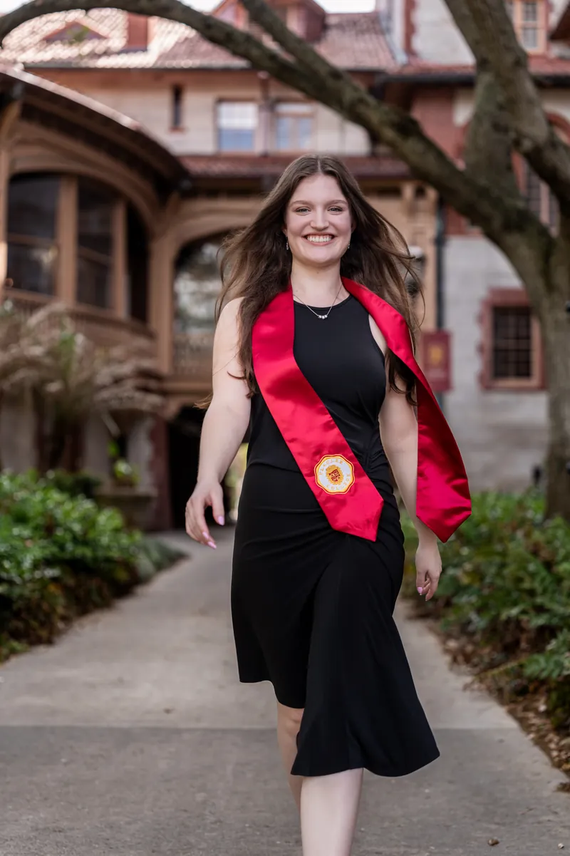 Olivia smiles walking toward the camera in a black sleeveless dress with a red floral lei, framed by green shrubs and arches