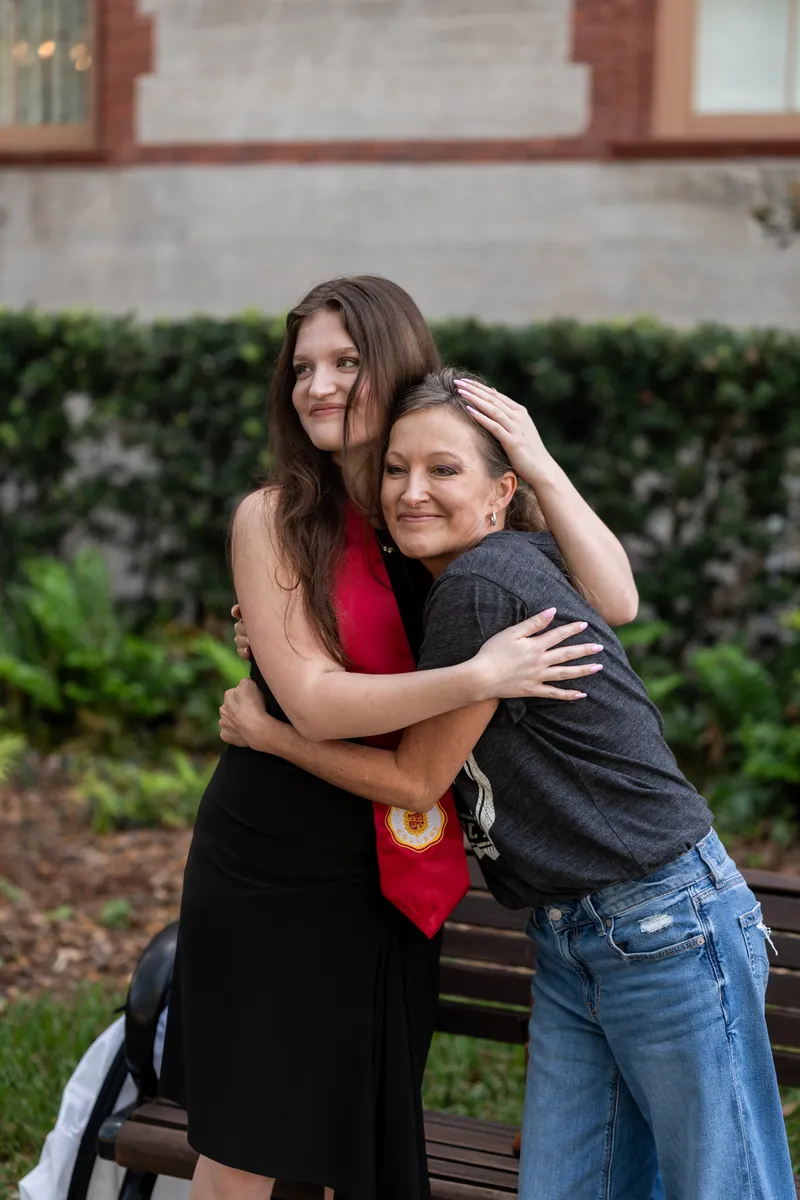 Two young women embrace on a bench at ivy-covered wall, one in a black dress with red scarf, the other in jeans