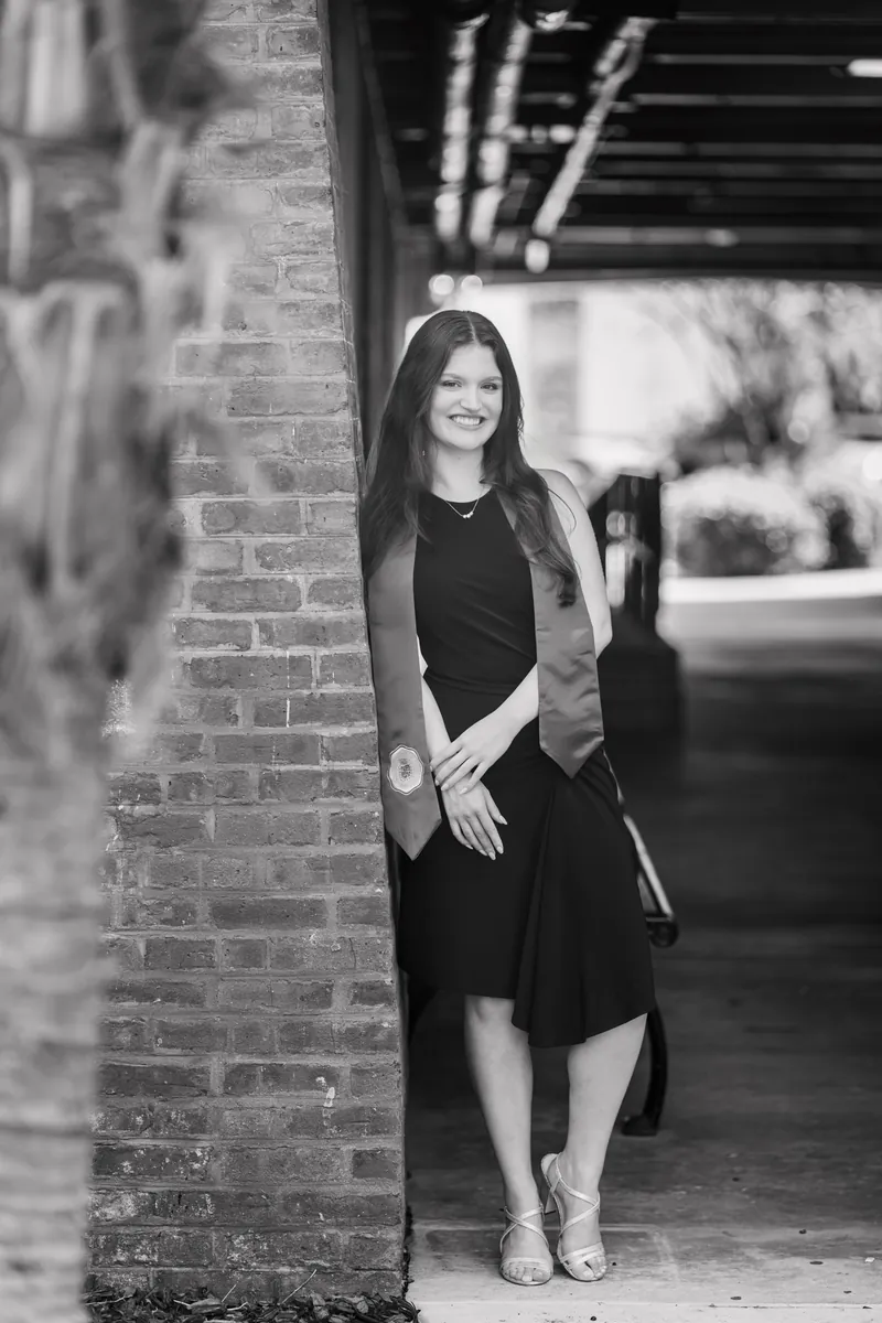 Black and white portrait of Olivia leaning against a brick wall in a black sleeveless dress under an overhang